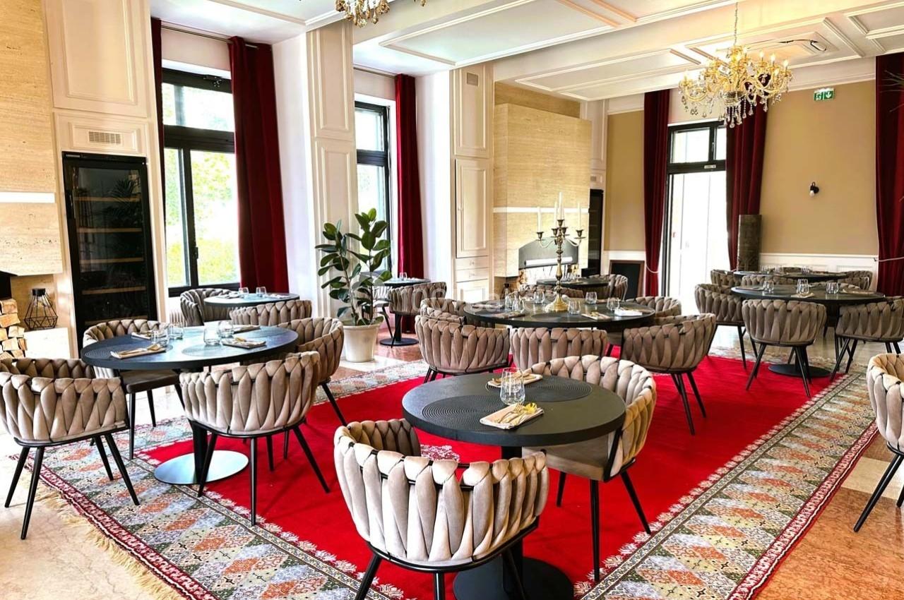 Salle de restaurant élégante avec tables rondes noires, chaises capitonnées, tapis rouge et grandes fenêtres lumineuses.
