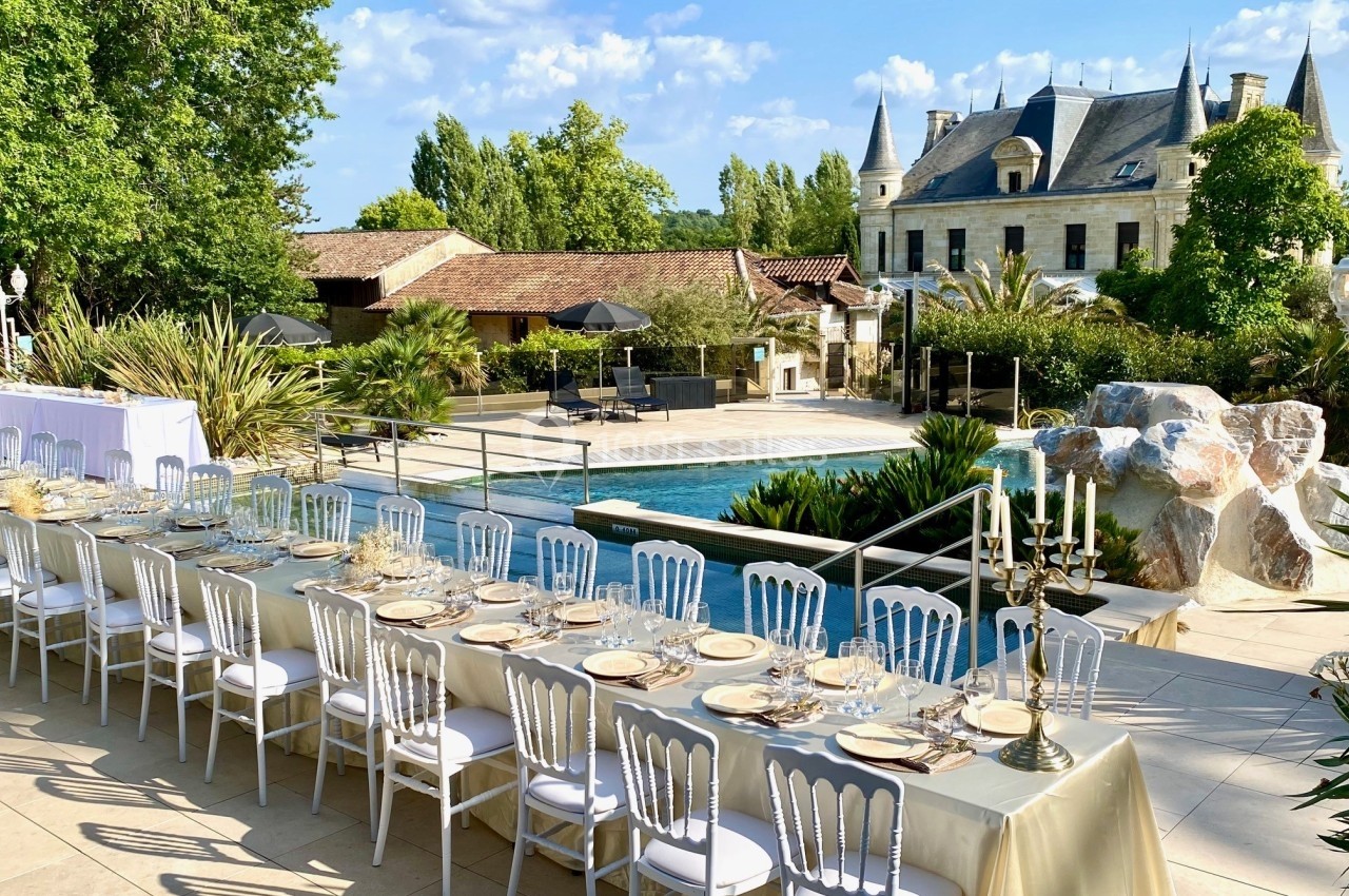 Grande table dressée en extérieur près d'une piscine, entourée de végétation et d'un bâtiment de style château.