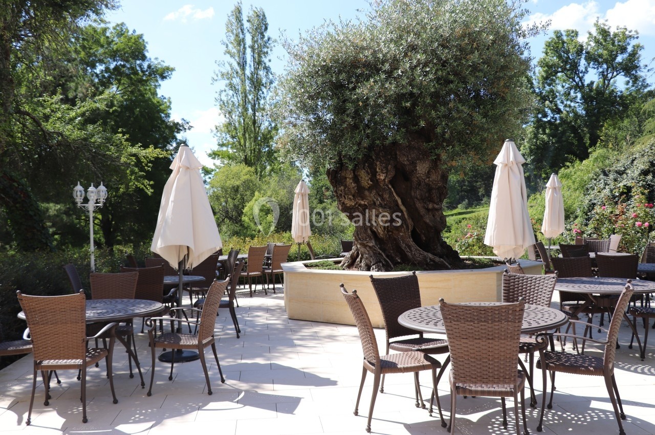Terrasse avec tables et chaises en osier autour d'un grand olivier, entourée de verdure et de parasols beige.