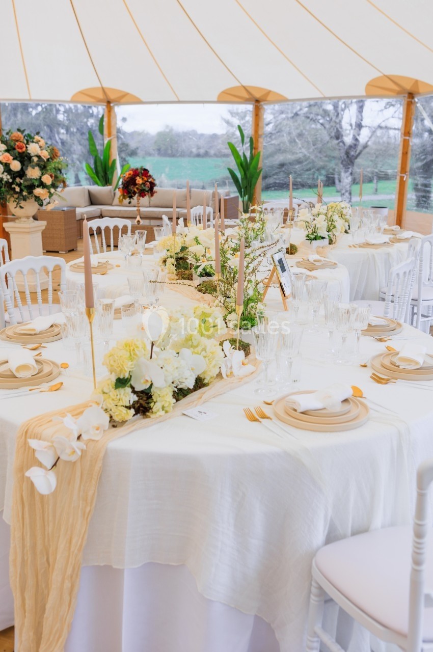Table dressée pour un événement, décorée de fleurs blanches et beige, avec vaisselle élégante sous une tente lumineuse.