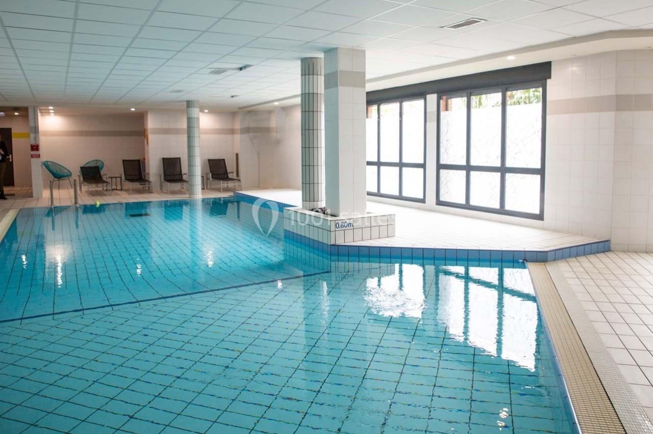Piscine intérieure avec carrelage bleu, entourée de chaises longues et éclairée par des fenêtres laissant entrer la lumière…