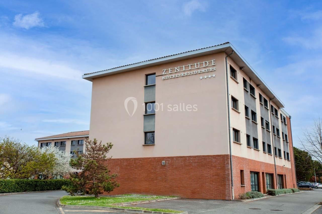 Façade d'un bâtiment moderne à trois étages avec des murs beige et briques, entouré de végétation sous un ciel bleu.