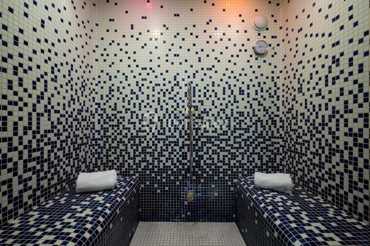 Salle de hammam avec murs et bancs en mosaïque bleue, équipée d'une douche murale et de serviettes pliées.