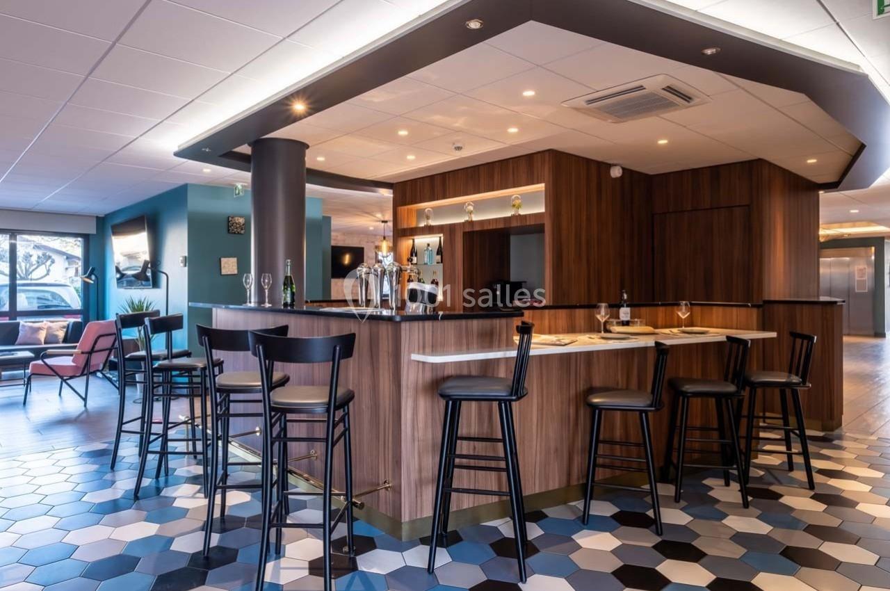 Bar moderne avec comptoir en bois, tabourets noirs, sol en carrelage hexagonal et ambiance lumineuse chaleureuse.