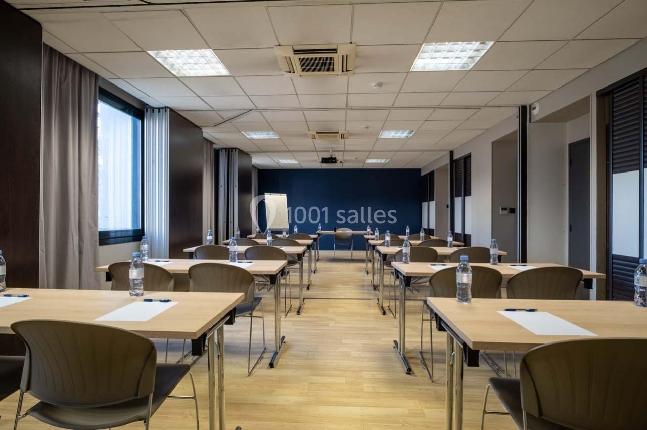 Salle de réunion lumineuse avec des tables disposées en rangées, chaises, bouteilles d'eau et tableau de présentation.