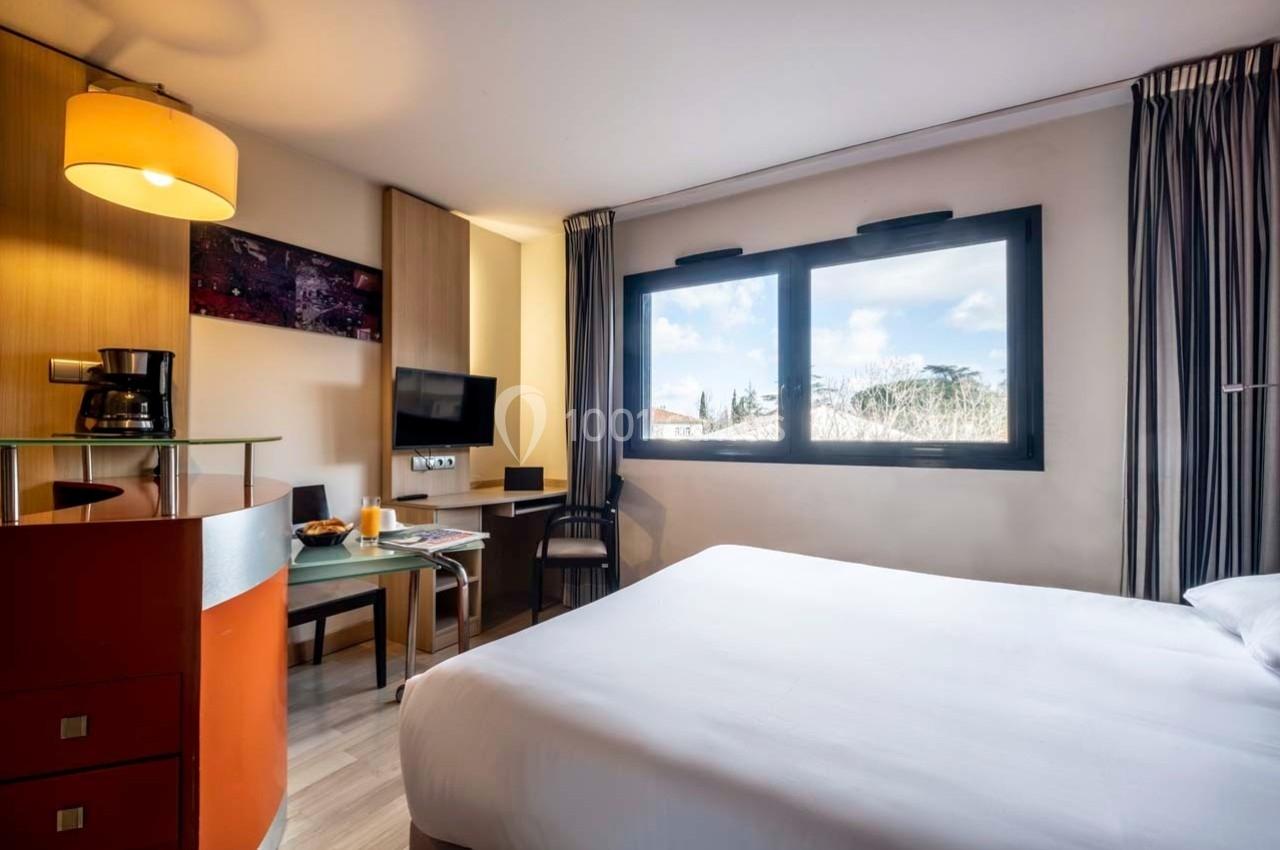 Chambre lumineuse avec lit double, bureau, télévision, machine à café et grande fenêtre offrant une vue extérieure.