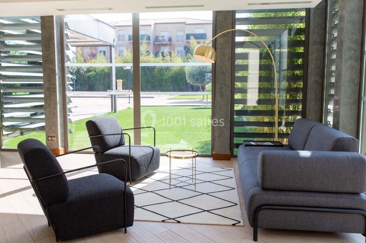 Salon moderne avec fauteuils gris, canapé assorti, lampe dorée et vue sur un jardin à travers de grandes baies vitrées.