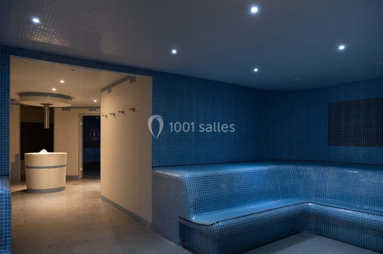 Salle de hammam avec bancs carrelés bleus, éclairage tamisé et vue sur un espace de réception en arrière-plan.
