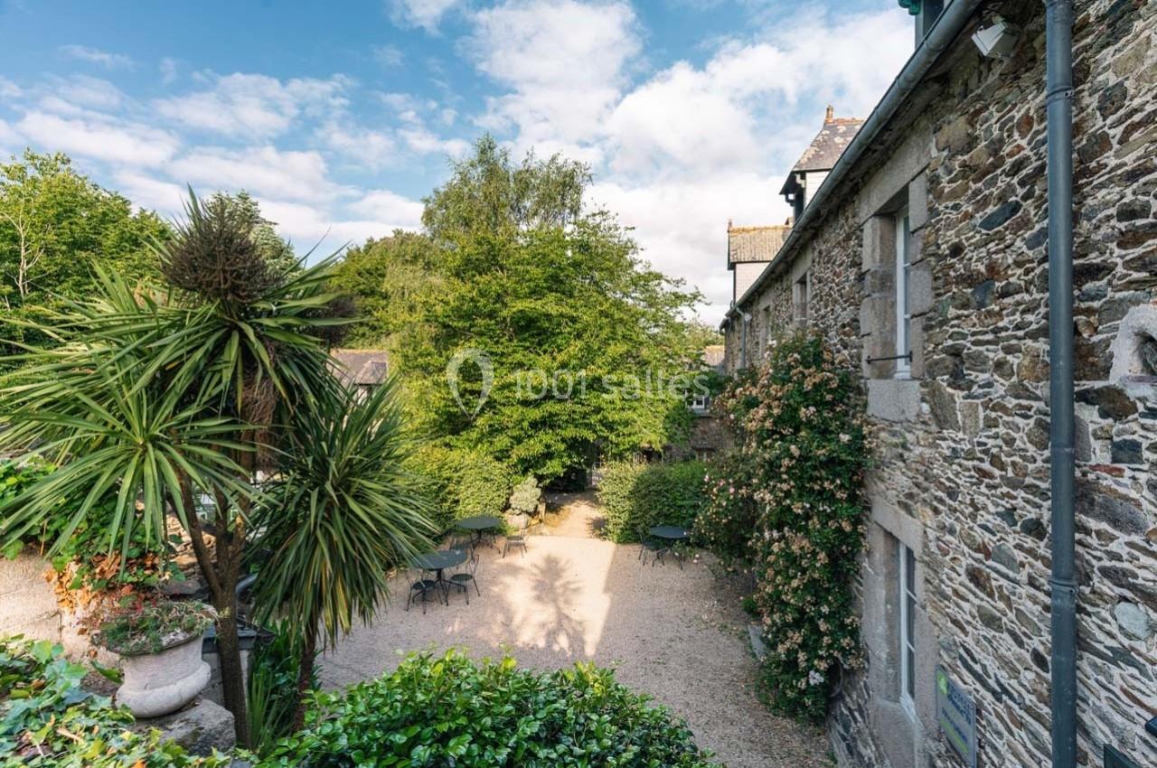 Cour extérieure avec tables et chaises entourées de végétation luxuriante et d'un bâtiment en pierre sous un ciel dégagé.