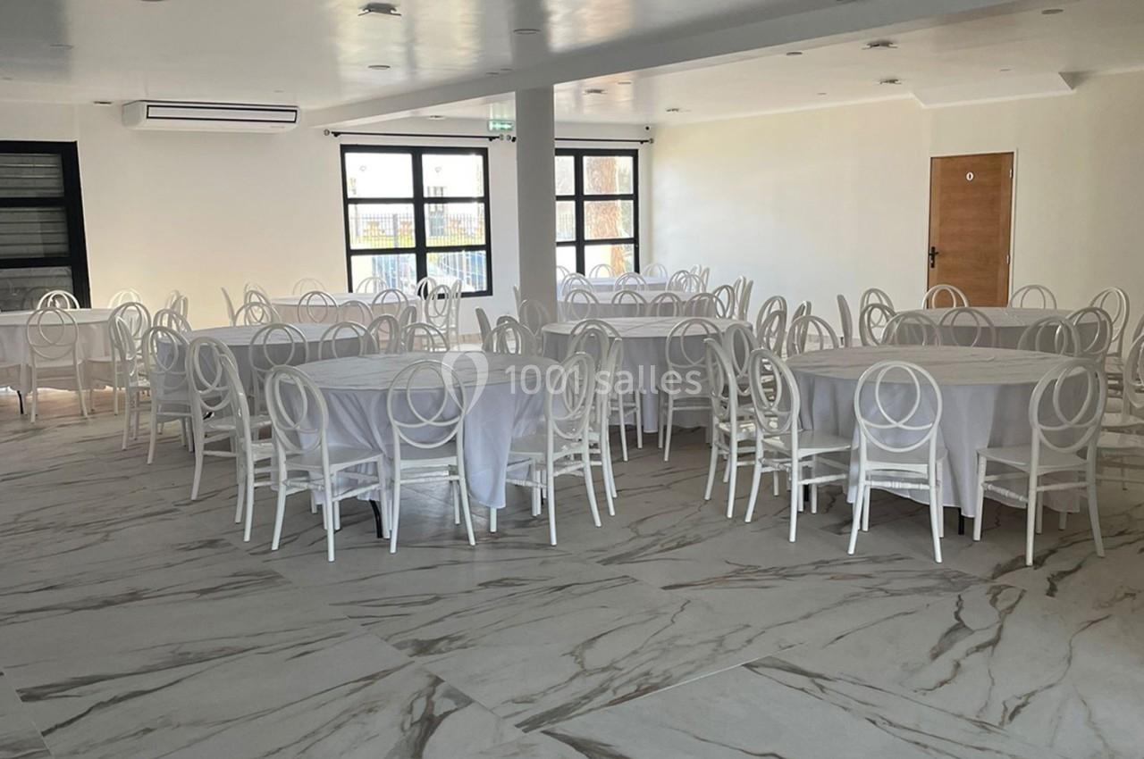 Salle lumineuse avec tables rondes recouvertes de nappes blanches et chaises blanches, sol en marbre clair.