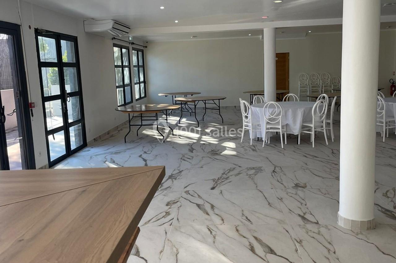 Salle lumineuse avec sol en marbre, tables rondes et rectangulaires, chaises blanches, grandes fenêtres et murs clairs.