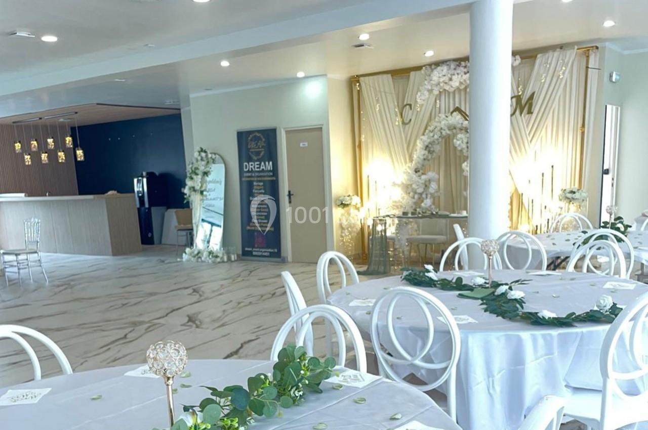 Salle de réception lumineuse avec tables rondes décorées de nappes blanches et guirlandes de feuillage.