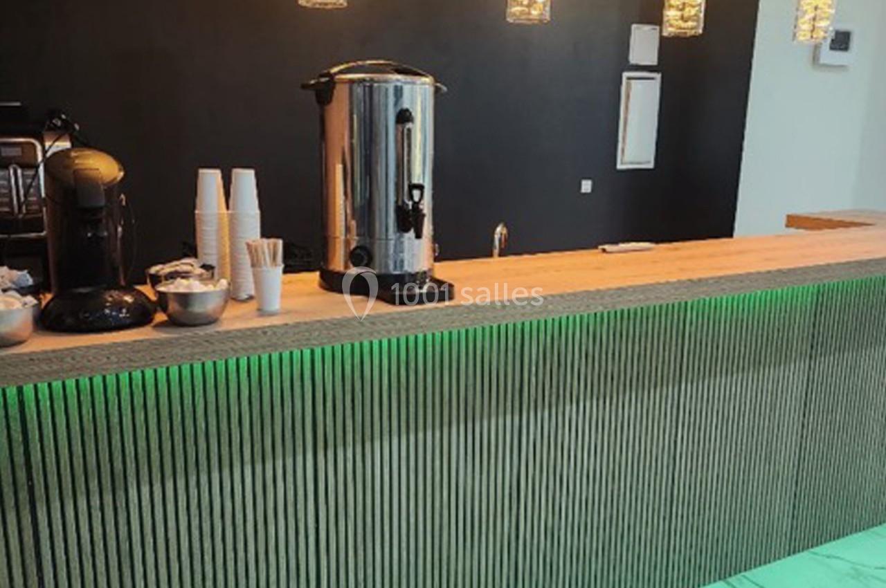 Comptoir en bois avec machine à café, distributeur d'eau chaude, gobelets et éclairage suspendu dans un espace moderne.