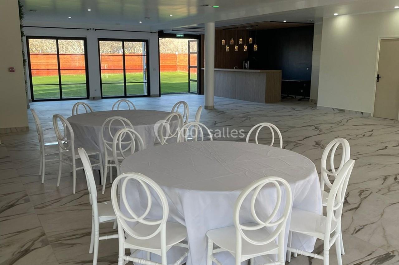 Salle lumineuse avec tables rondes couvertes de nappes blanches, chaises blanches et vue sur un espace extérieur.