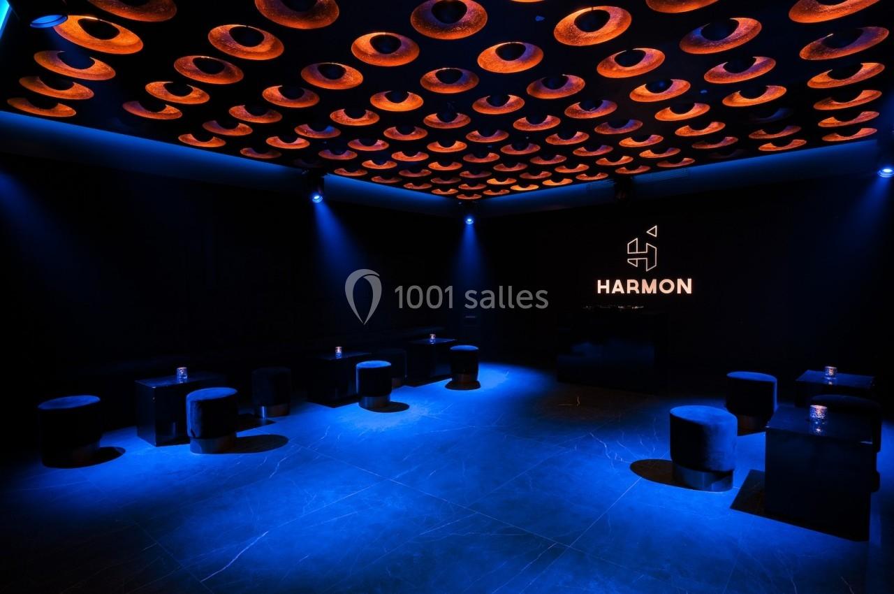 Salle sombre avec un éclairage bleu, plafond orné de cercles lumineux orange et logo ’Harmon’ illuminé sur un mur.