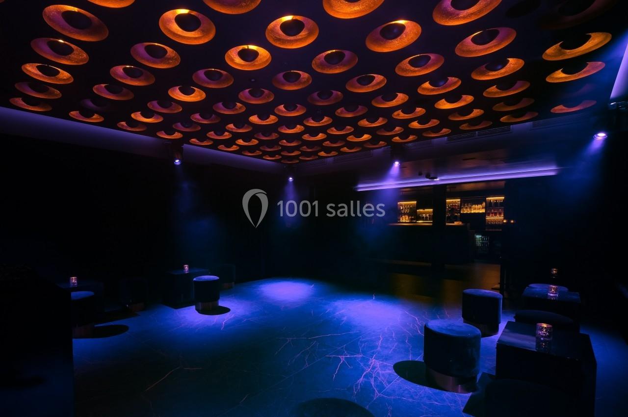 Salle sombre avec un plafond décoratif circulaire illuminé, des tabourets noirs et un bar éclairé en arrière-plan.
