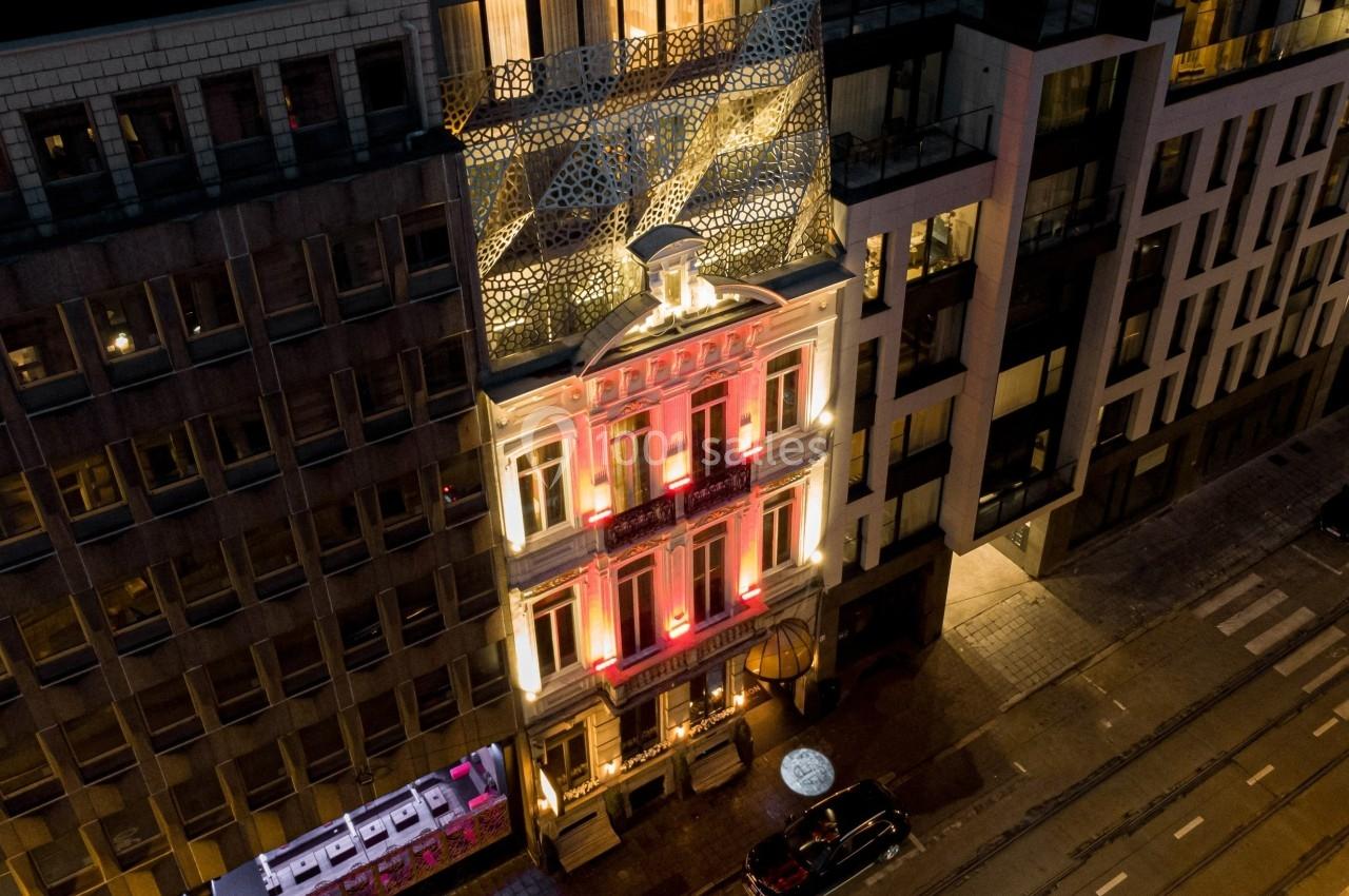 Façade illuminée d'un bâtiment ancien entouré d'immeubles modernes, vue de nuit depuis une perspective aérienne.