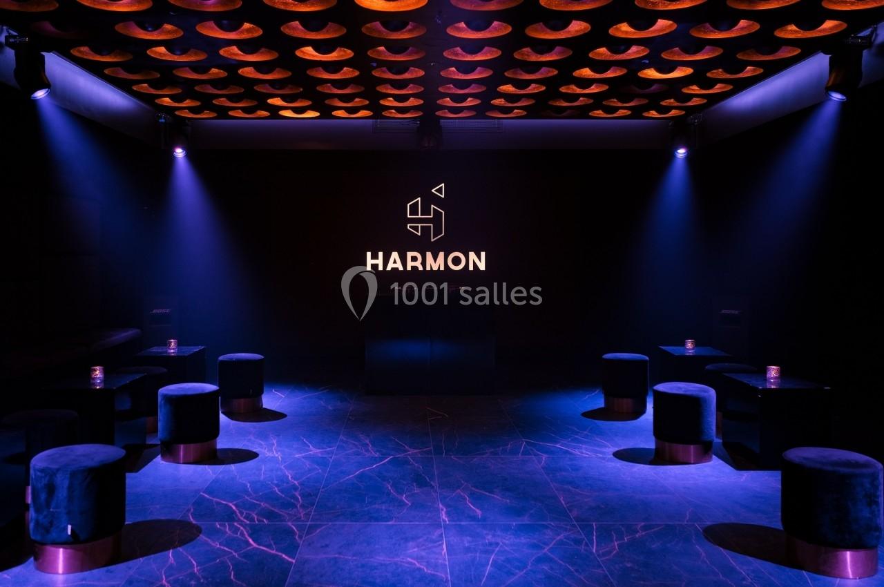 Salle sombre avec un éclairage violet, des tabourets noirs et un logo lumineux ’HARMON’ sur un mur noir.