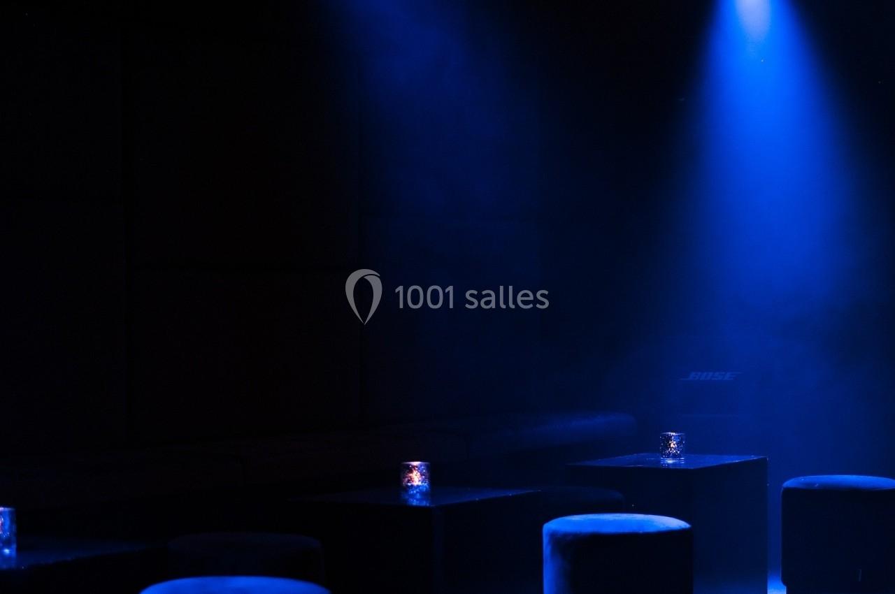 Salle sombre avec éclairage bleu, tabourets ronds noirs et petites bougies sur des tables basses.