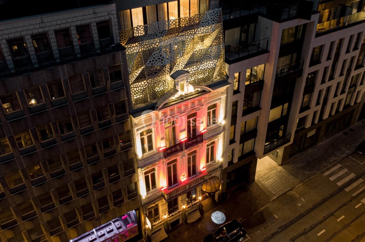 Vue nocturne d'un bâtiment illuminé avec une façade moderne et un toit décoratif, entouré d'autres immeubles urbains.