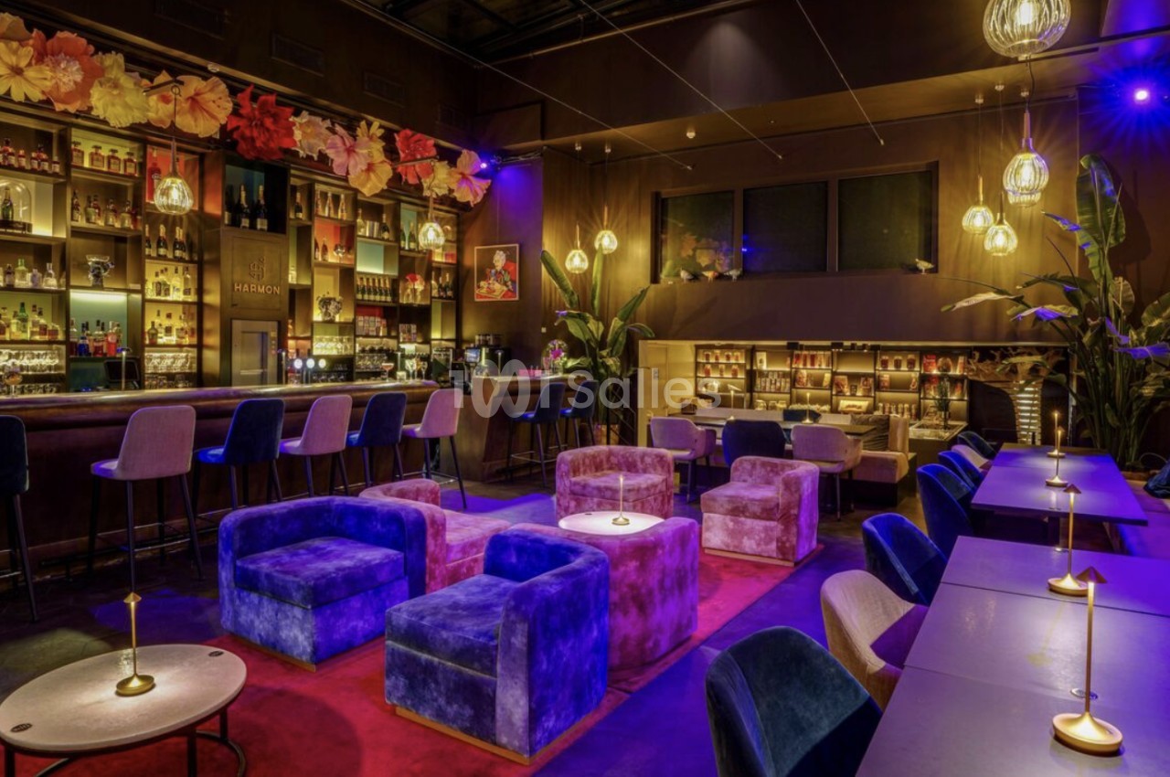 Salle de bar intérieure à l'éclairage violet et ambré, avec un comptoir à tabourets roses et bleus, des fauteuils en velours et des tables hautes dressées. Let me check length: roughly 150 characters, too long. Need ≤125. Salle de bar à l'éclairage