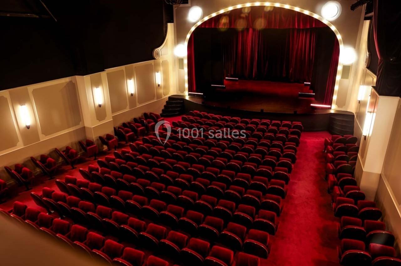 Salle de théâtre avec sièges rouges alignés face à une scène éclairée par des rideaux et des lumières.