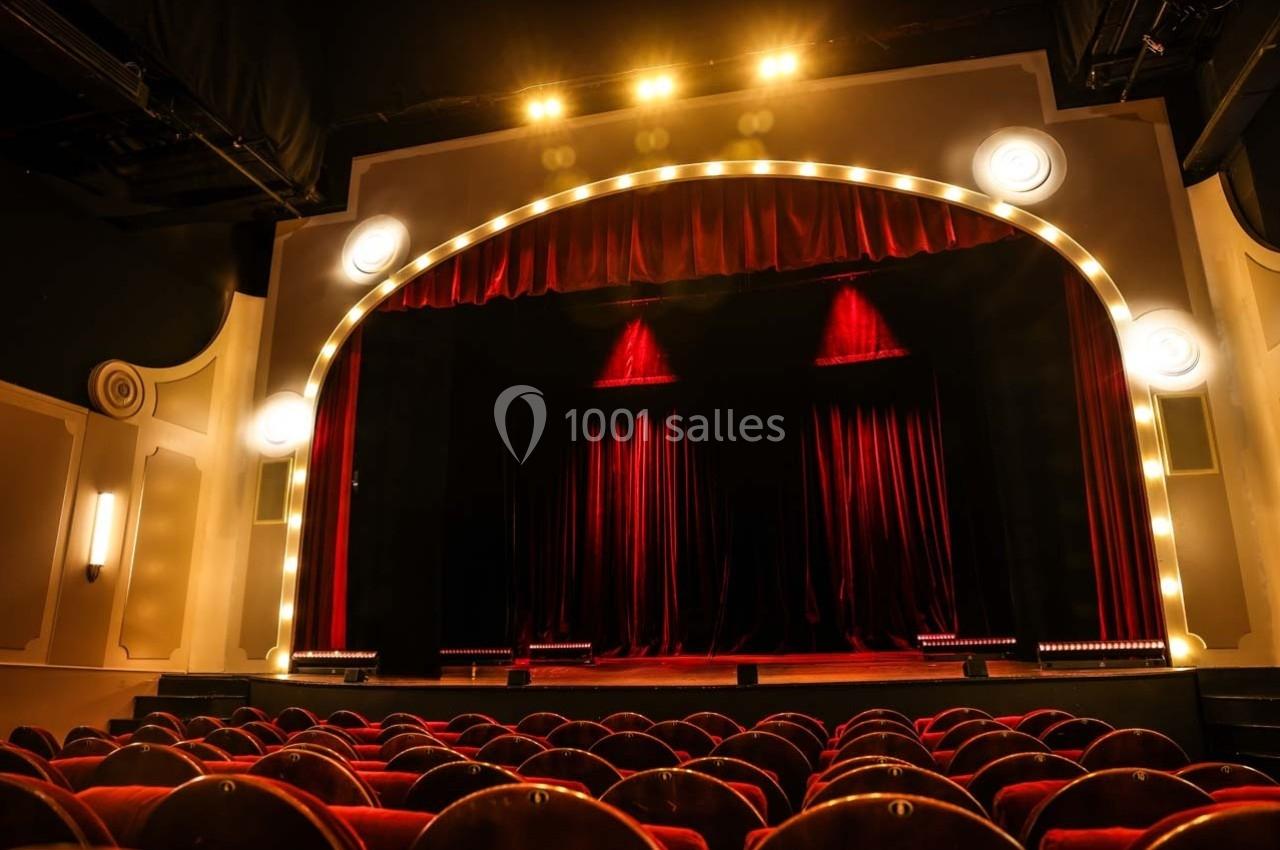 Salle de théâtre avec rideaux rouges fermés, éclairée par des projecteurs et entourée de sièges en velours rouge.