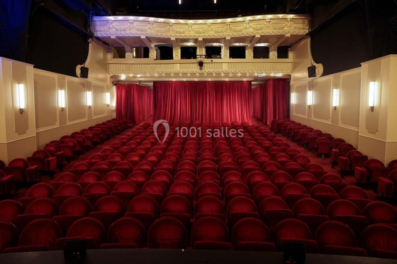 Salle de théâtre vide avec des rangées de fauteuils rouges et une scène fermée par un rideau rouge.