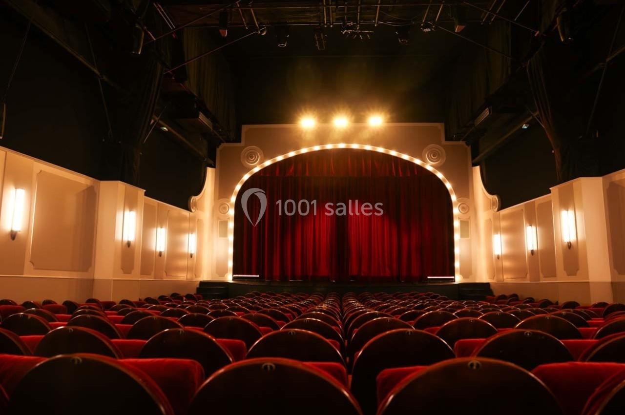 Salle de théâtre vide avec rangées de sièges rouges et une scène éclairée par des projecteurs.