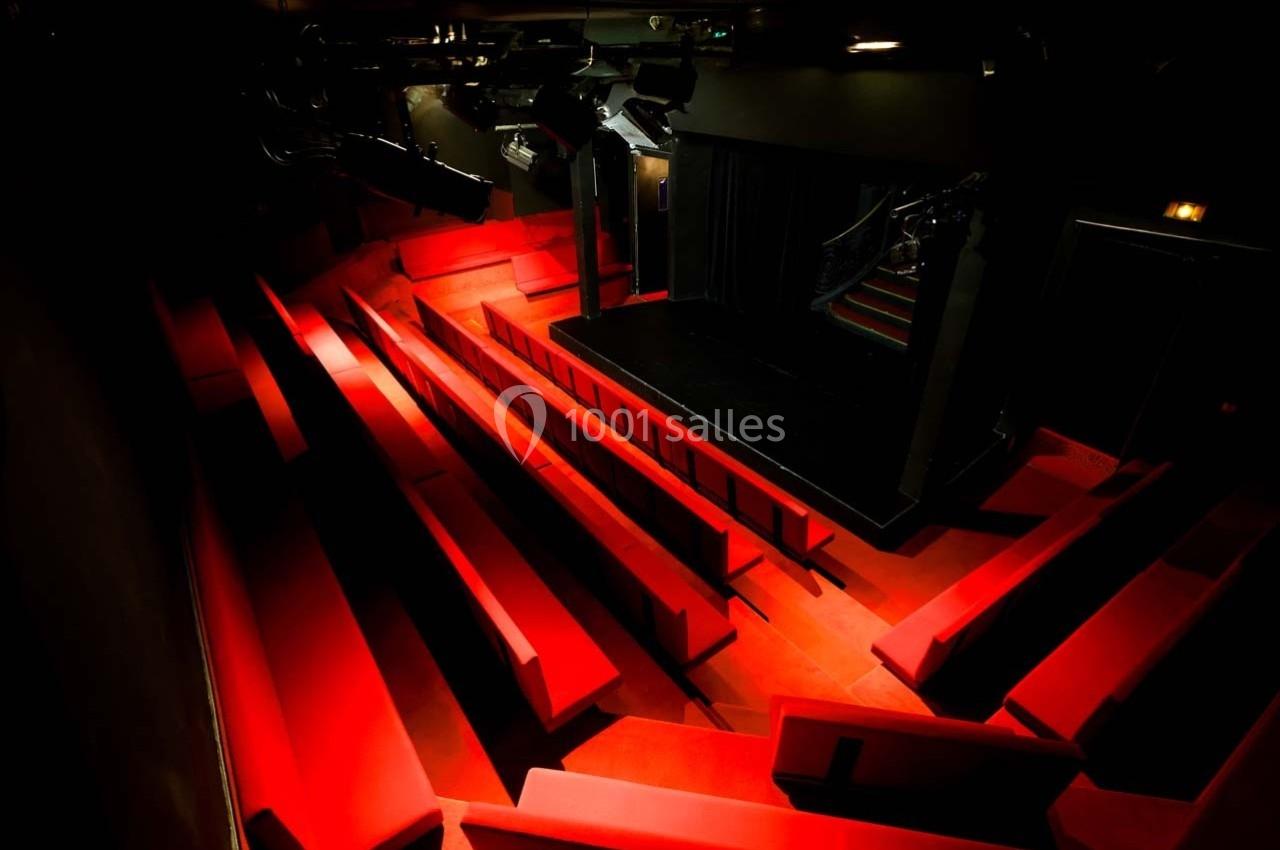 Salle de théâtre vide avec sièges rouges en gradins et une scène éclairée par des projecteurs.