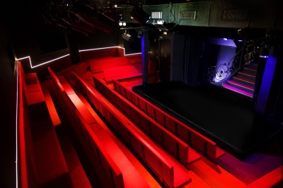 Salle avec moquette rouge, table en bois clair, chaises rouges et miroir éclairé par des ampoules.