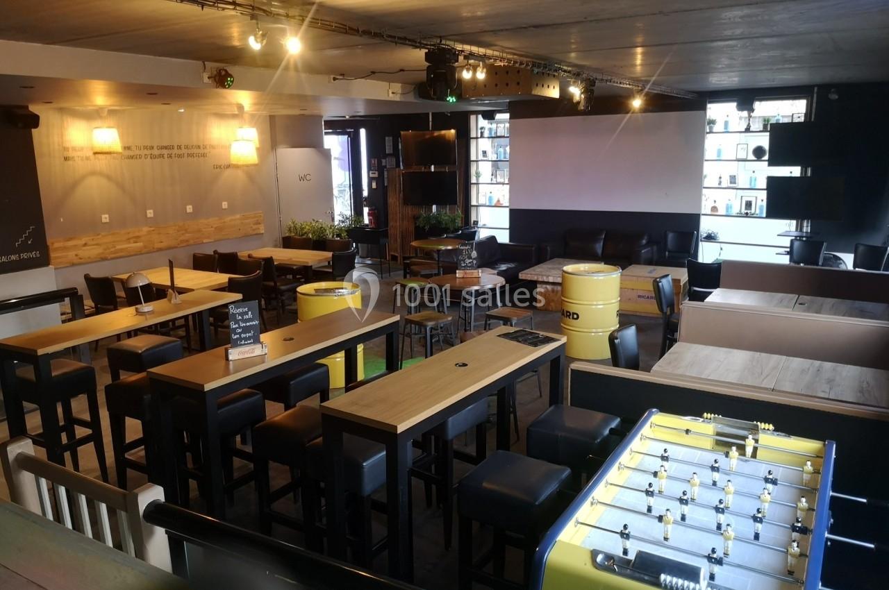 Salle de bar moderne avec tables hautes, tabourets, baby-foot jaune et éclairage chaleureux.