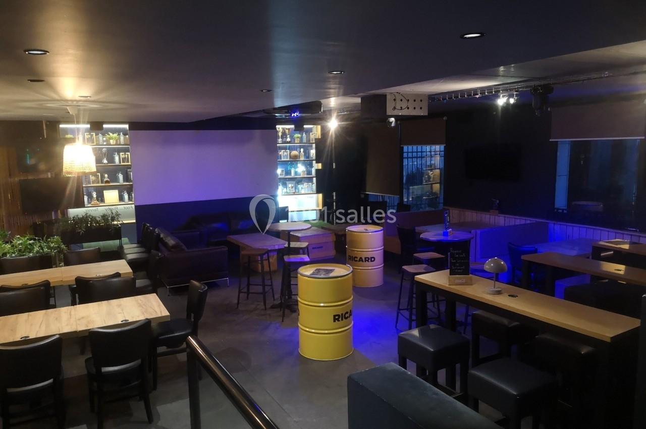 Salle de bar avec tables en bois, tonneaux jaunes en guise de tables, éclairage tamisé et étagères illuminées.