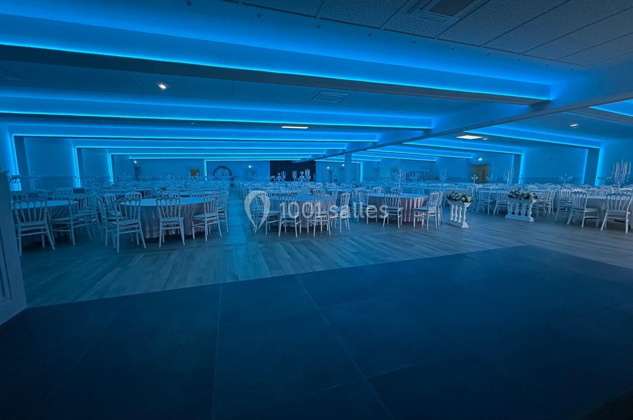 Salle de réception spacieuse avec tables et chaises blanches, éclairée par une lumière bleue tamisée.