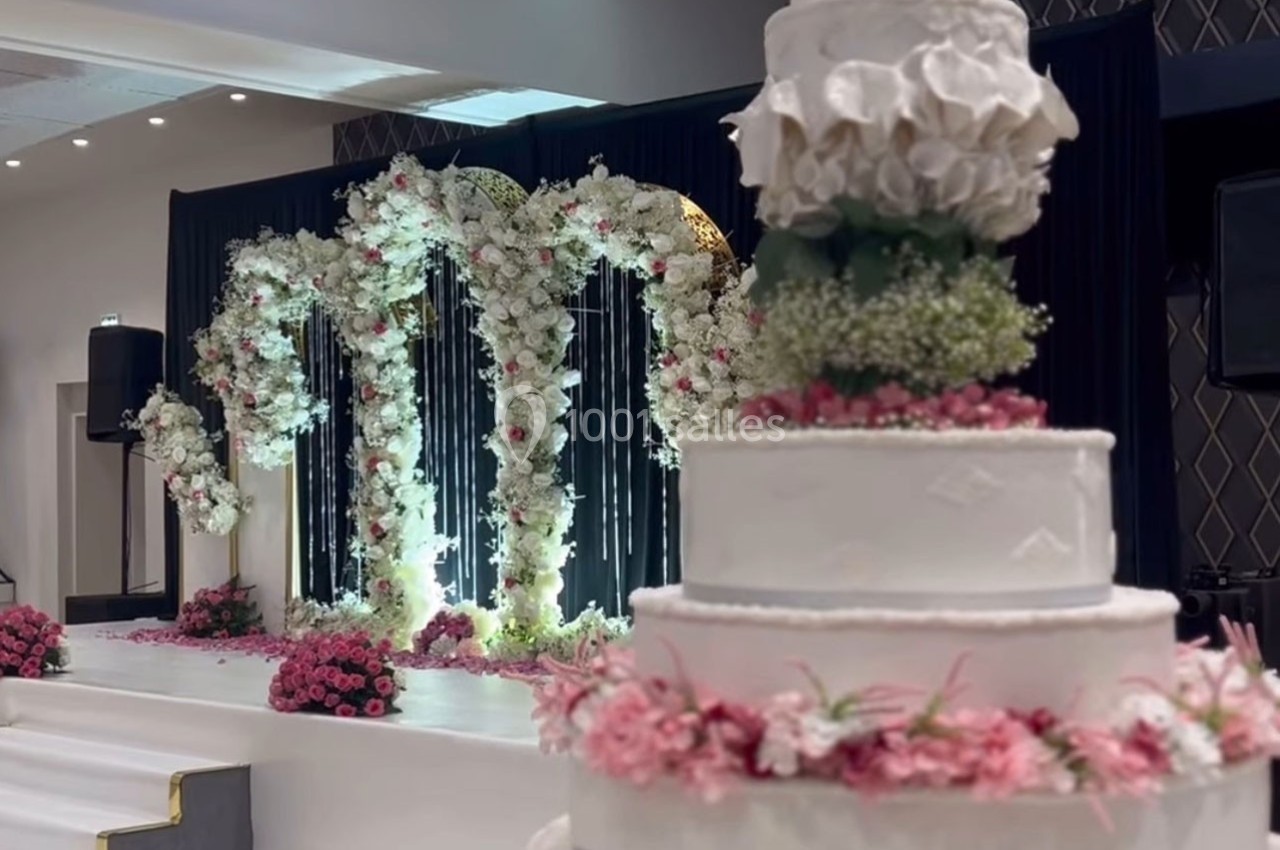 Décor de mariage avec arche florale blanche et rose en arrière-plan et gâteau de mariage décoré au premier plan.
