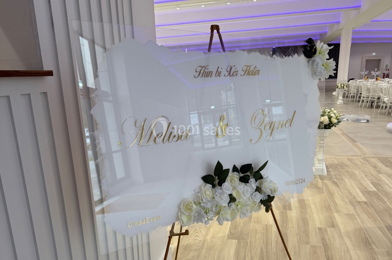 Panneau de mariage décoratif avec des fleurs blanches et des prénoms dorés, placé dans une salle lumineuse.