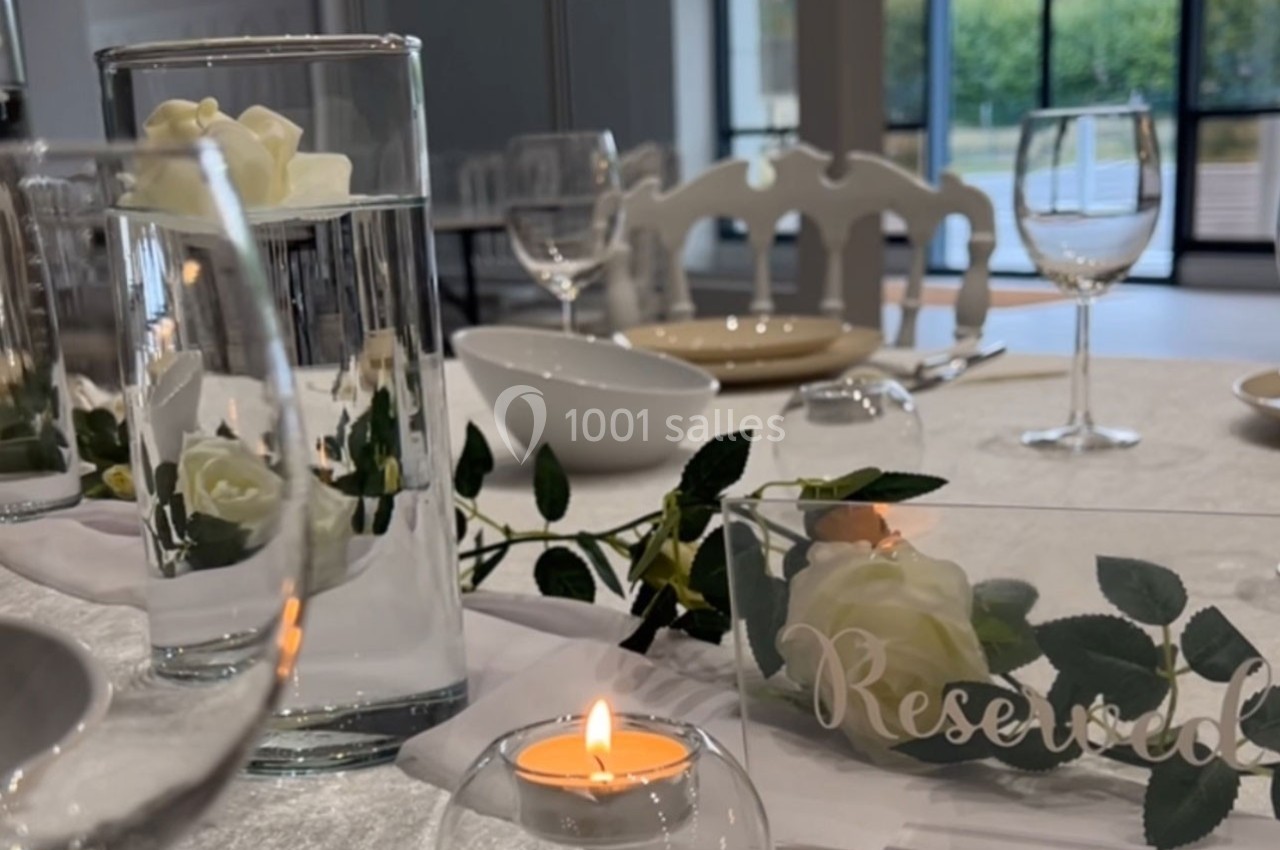 Table décorée pour un événement avec bougies, roses blanches, verres et assiettes, près de grandes fenêtres.