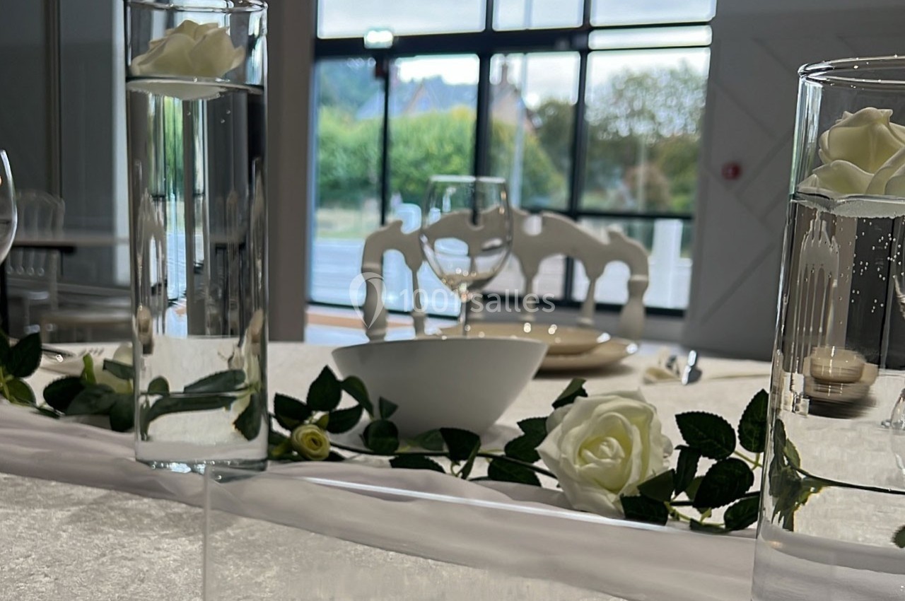 Décoration de table élégante avec roses blanches, verres et vaisselle, devant une grande baie vitrée lumineuse.