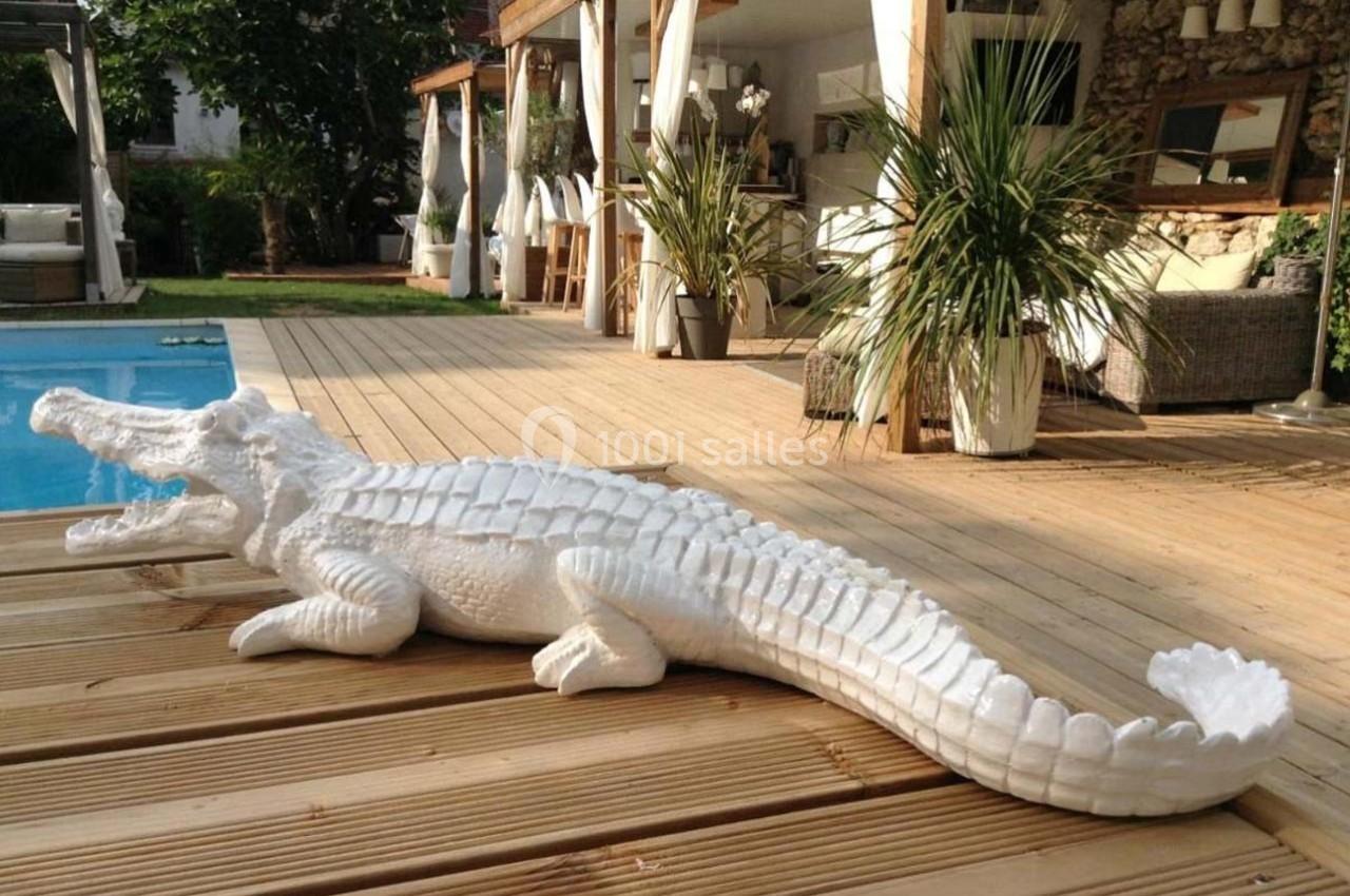 Statue blanche de crocodile posée sur une terrasse en bois près d'une piscine, entourée de plantes et mobilier extérieur.