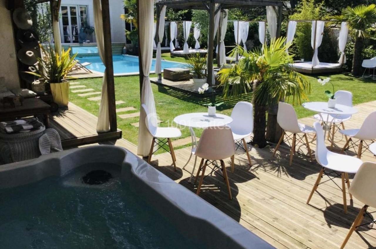 Terrasse en bois avec jacuzzi, tables et chaises blanches, vue sur un jardin avec piscine et lits à baldaquin.