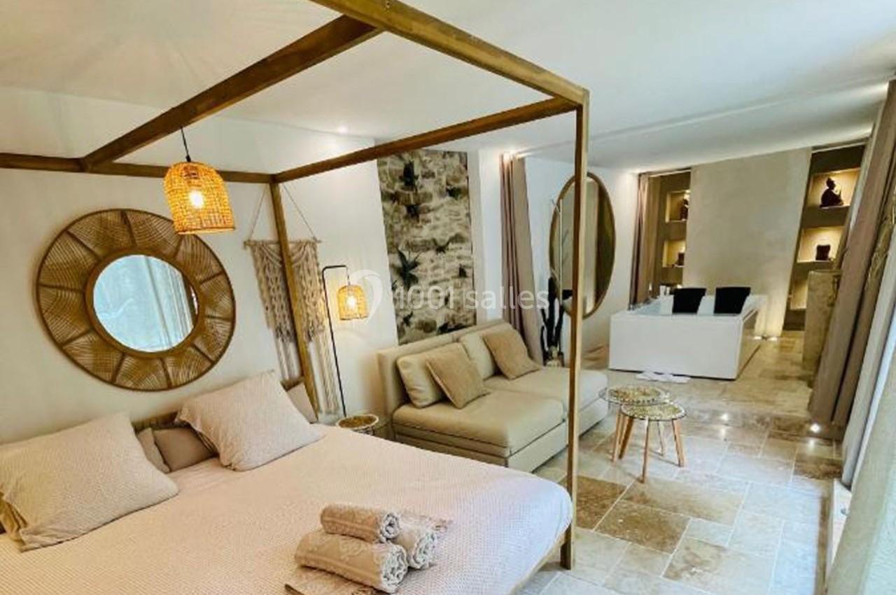 Chambre lumineuse avec lit à baldaquin, canapé beige, décoration naturelle et salle de bain ouverte en arrière-plan.