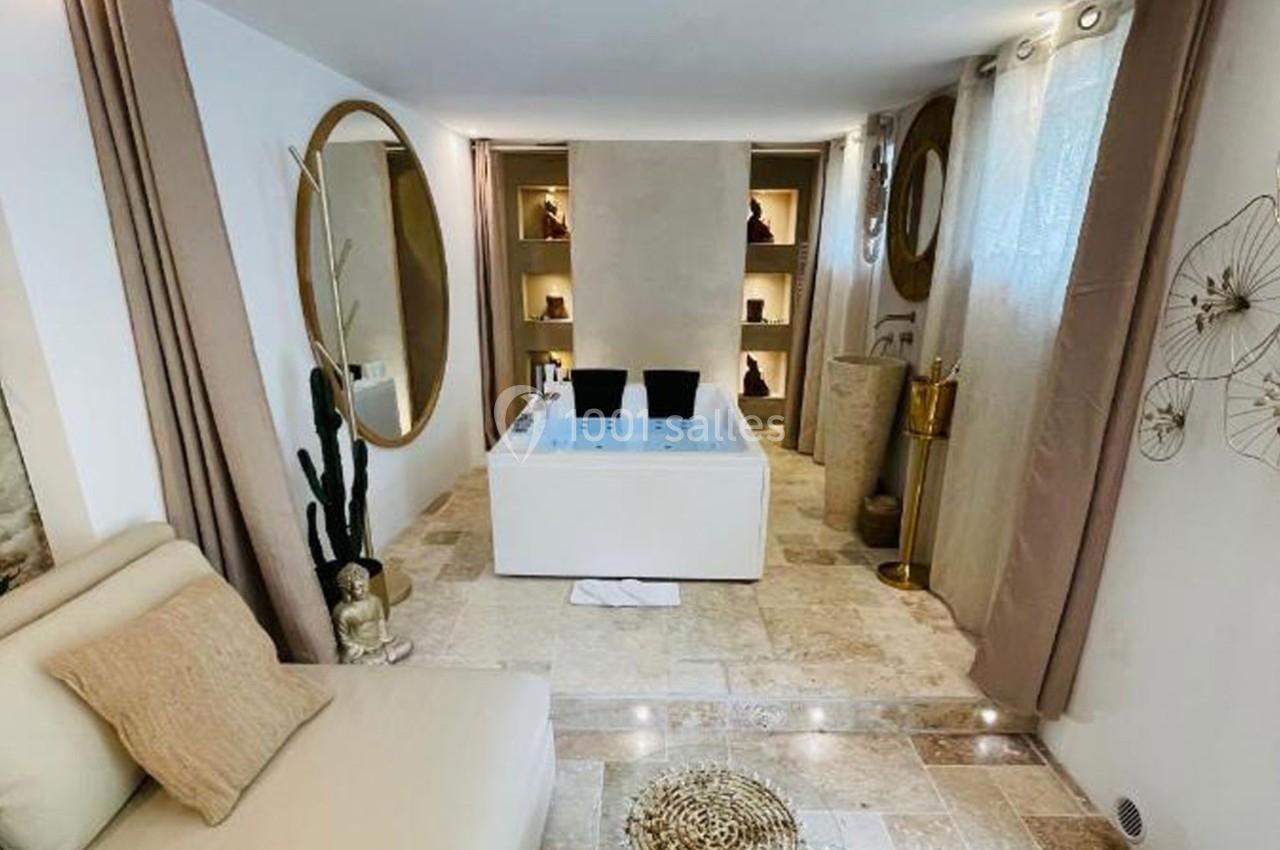 Salle de bain lumineuse avec baignoire centrale, fauteuil beige, miroir rond et décorations naturelles.