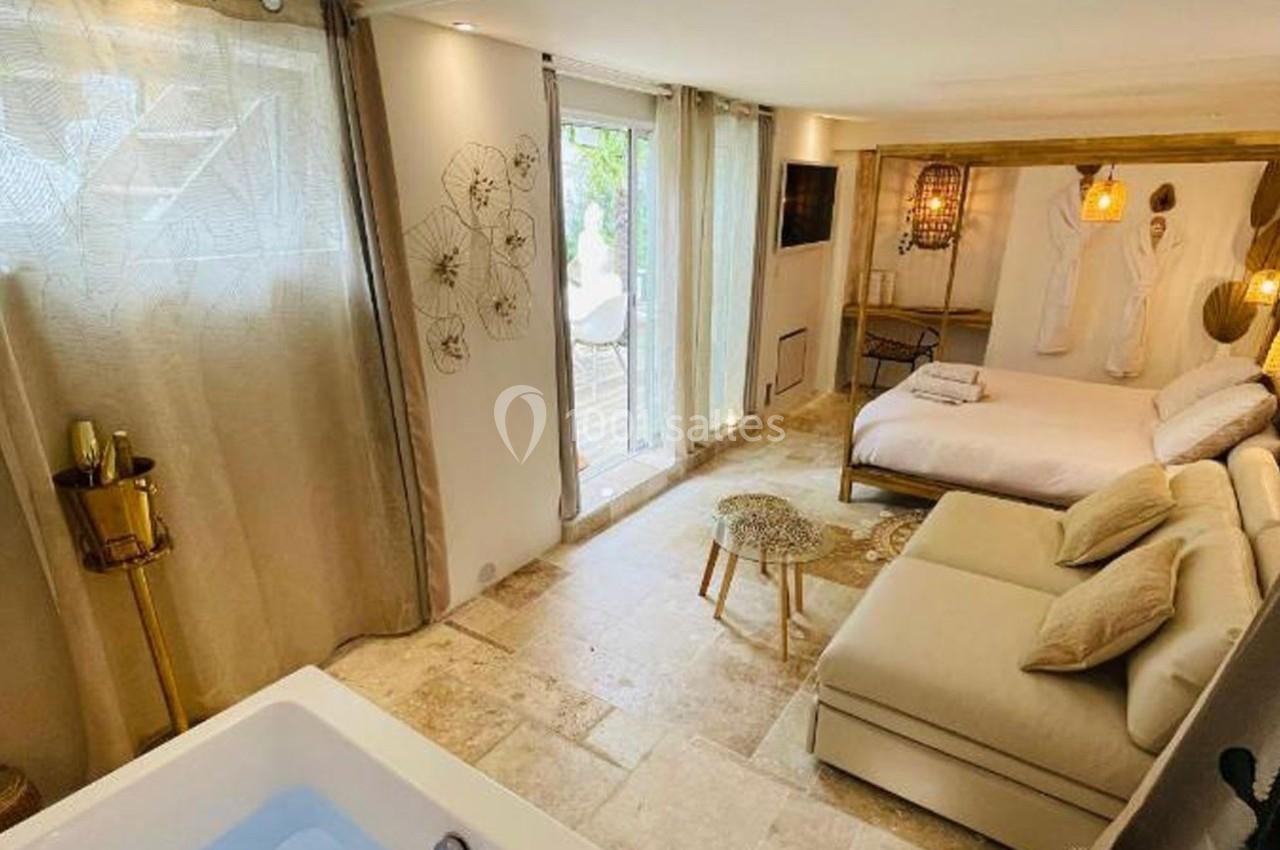 Chambre lumineuse avec lit double, canapé, jacuzzi, décor naturel et accès à une terrasse par une baie vitrée.