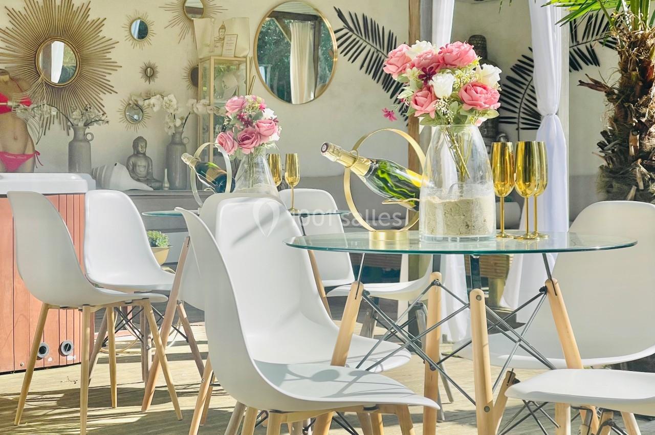 Table en verre entourée de chaises blanches, décorée de fleurs roses, verres dorés et bouteille de champagne, en extérieur.