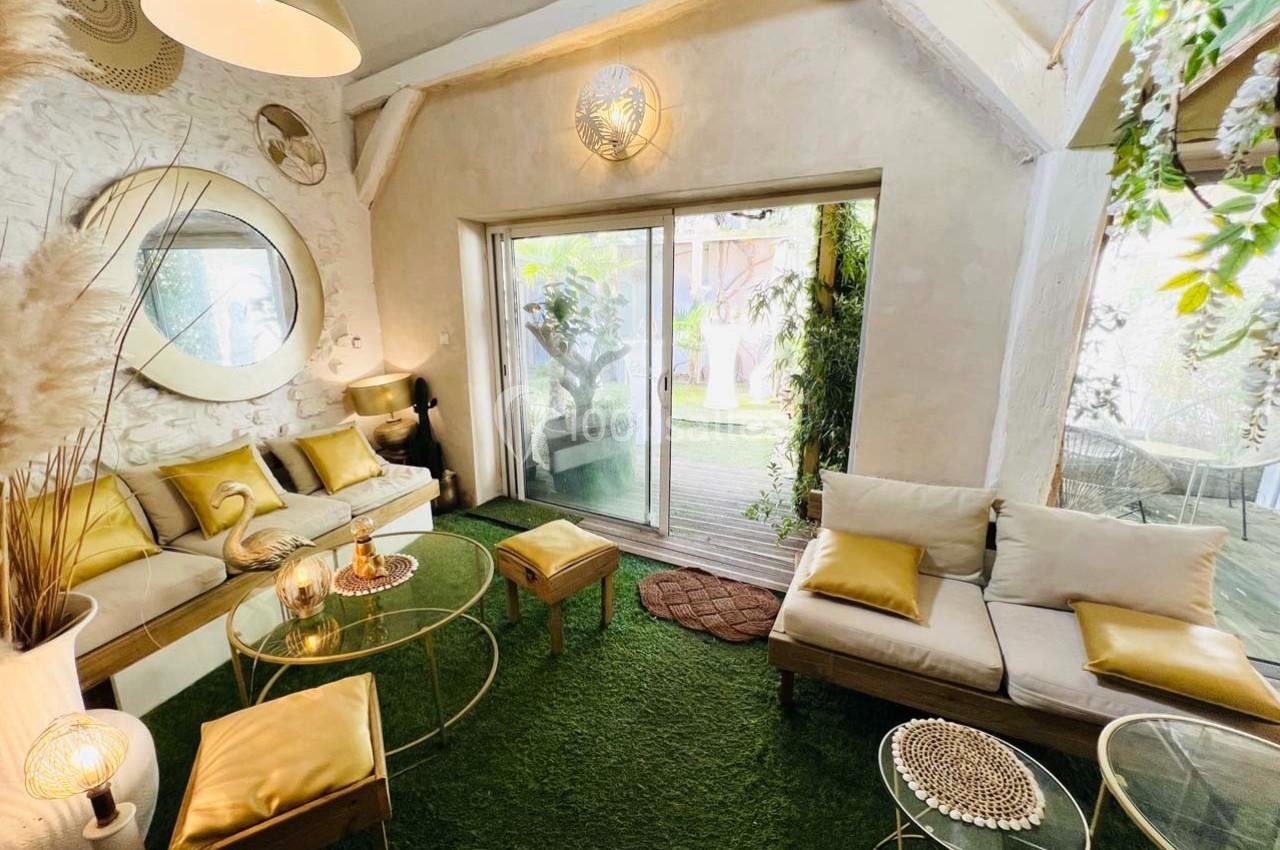 Salon lumineux avec canapés clairs, coussins dorés, tables en verre et vue sur une terrasse végétalisée par une baie vitrée.