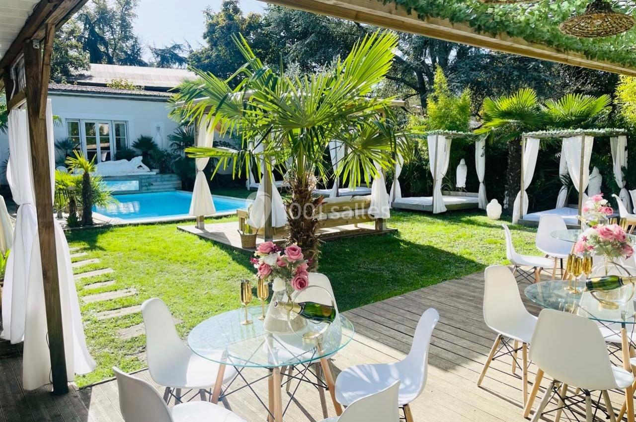 Jardin avec piscine, tables en verre, chaises blanches et décoration florale, entouré de végétation tropicale.