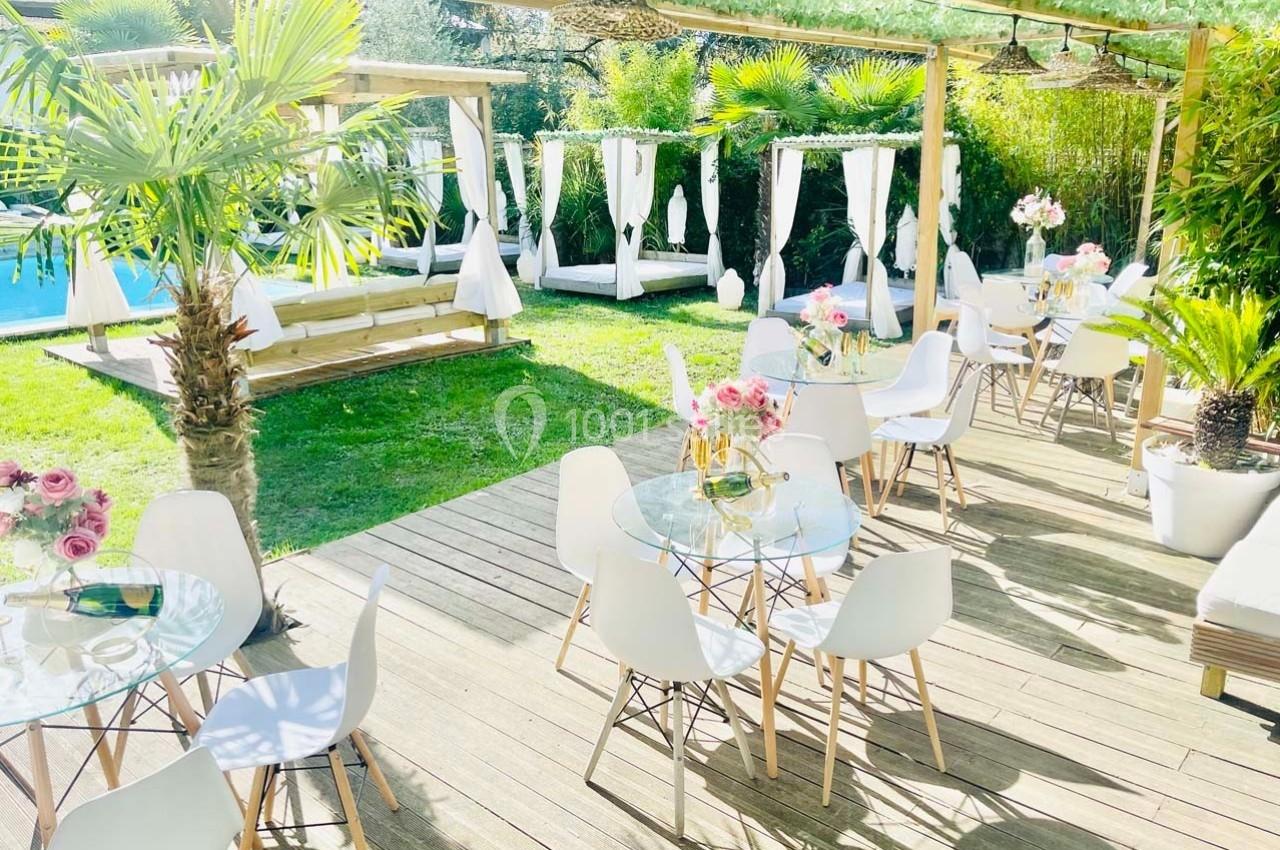 Terrasse ensoleillée avec tables en verre, chaises blanches, plantes exotiques et vue sur une piscine entourée de verdure.