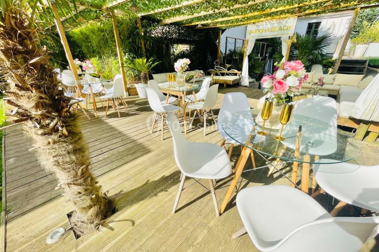 Terrasse ensoleillée avec tables en verre, chaises blanches, plantes exotiques et décorations florales.