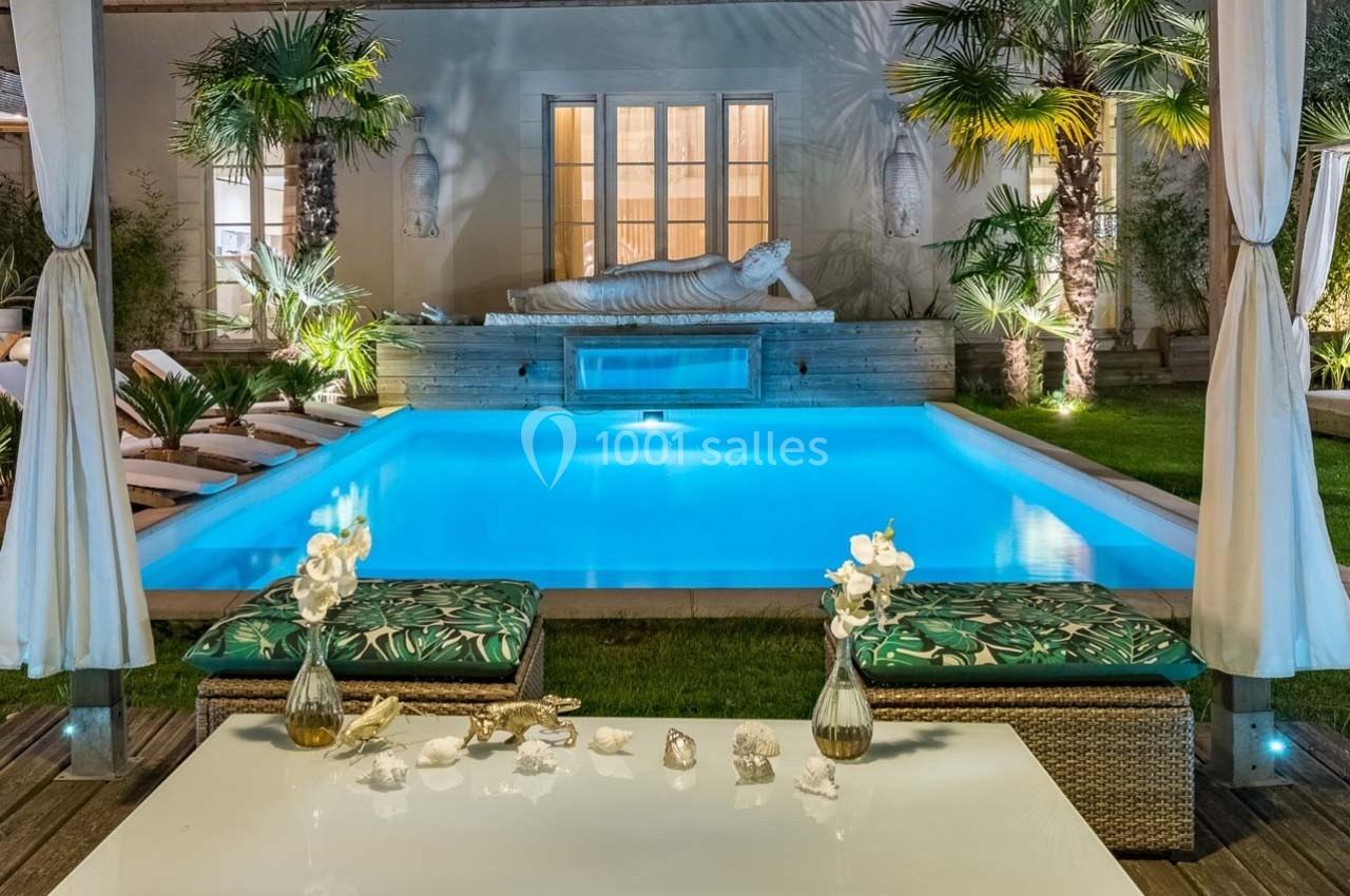 Piscine éclairée entourée de palmiers et de chaises longues, avec une statue décorative en arrière-plan.