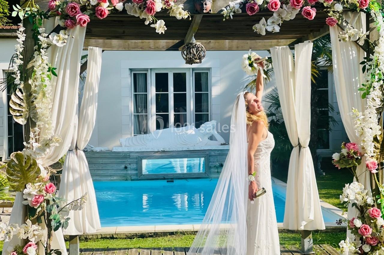 Une mariée en robe blanche lève un bouquet sous une pergola décorée de fleurs, devant une piscine et une statue.